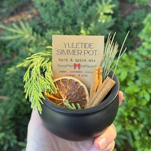 May include: A matte black cauldron filled with a Yuletide Simmer Pot herb and spice mix, featuring a dried orange slice, cinnamon sticks, and evergreen sprigs. A tan card displays the text: YULETIDE SIMMER POT herb & spice mix cedar, cinnamon, clove, orange, & pine.