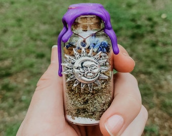 May include: A small glass bottle with a purple wax seal, filled with dried herbs and a silver sun and moon charm. The bottle is held in a hand.