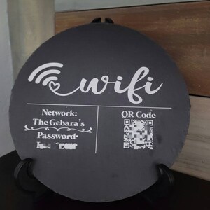 May include: Black slate sign with a white wifi symbol and the text "wifi". The sign also includes the text "Network: The Gebara's" and "Password:" with a blacked out password. A QR code is also included with the text "QR Code".