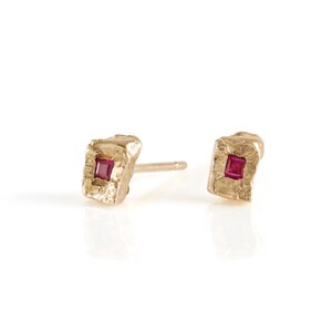 May include: A pair of gold earrings with a square ruby gemstone set in each. The earrings have a textured, hammered finish.
