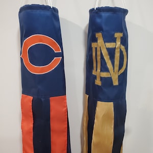 May include: Two blue fabric pennants with white strings. The pennant on the left has a red and white striped bottom and an orange "C" on the blue background. The pennant on the right has a gold and white striped bottom and a gold "ND" on the blue background.