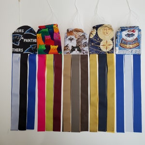 May include: Seven colorful fabric windsocks hanging from the ceiling. The windsocks are made of different fabrics and have various designs, including cats, a football team logo, a sun and moon design, and a snowman.
