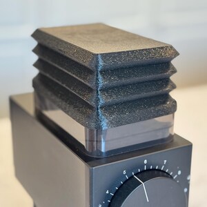 May include: Close-up of a black coffee grinder with a textured, ridged top. The grinder has a square base with a dial and numbers. The top section is a clear container. The overall design is modern and minimalist.