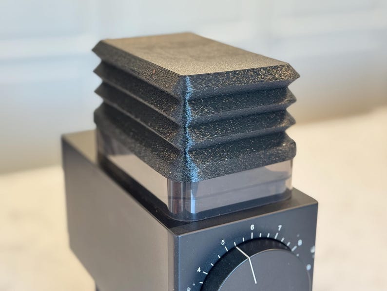 May include: Close-up of a black coffee grinder with a textured, ridged top. The grinder has a square base with a dial and numbers. The top section is a clear container. The overall design is modern and minimalist.