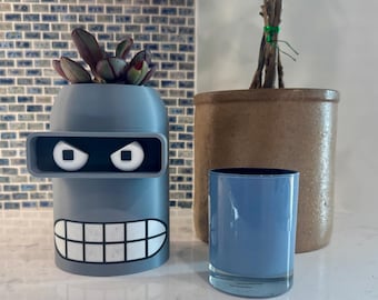 May include: A gray Bender-shaped planter with a white-toothed grin and square eyes, holding a succulent plant. Beside it, a blue glass and a brown ceramic pot with branches. A fun, decorative item for home decor.