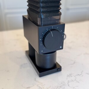 May include: A black electric coffee grinder with a textured top, a dial, and a cylindrical base. The grinder sits on a rectangular base and is designed for home use.