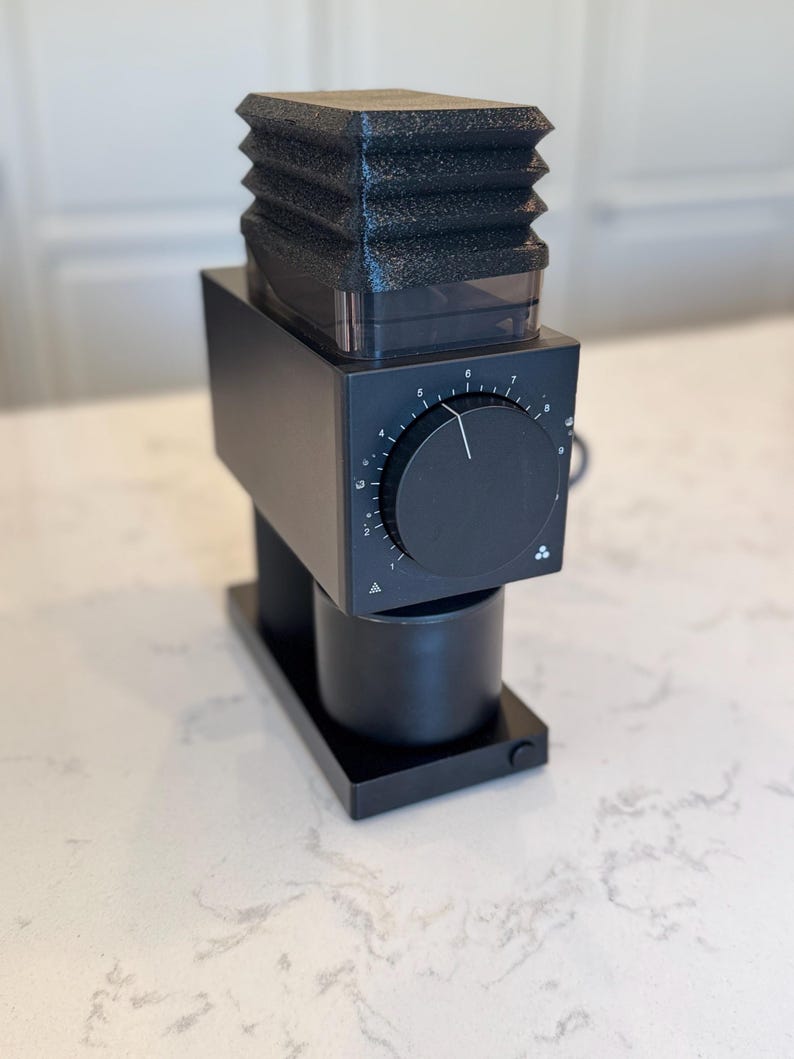 May include: A black electric coffee grinder with a textured top, a dial, and a cylindrical base. The grinder sits on a rectangular base and is designed for home use.