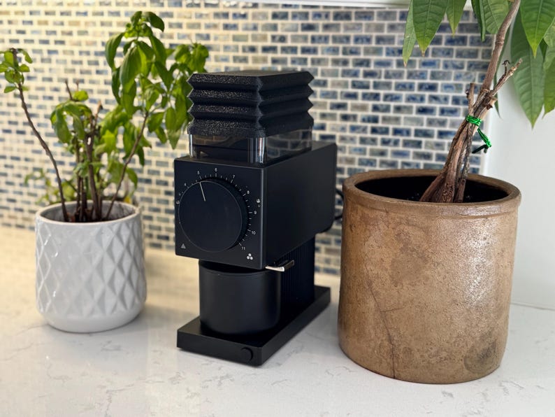 May include: A black electric coffee grinder with a dial and a small cup, next to a white patterned pot with a green plant and a brown pot with a plant. The grinder is on a white countertop with a blue and white tile backsplash.