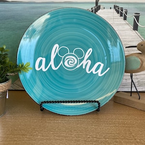 May include: A turquoise ceramic plate with a white "aloha" design, featuring a Mickey Mouse-inspired swirl. The plate has a spiral pattern and a dark brown rim. It is displayed on a black metal stand.
