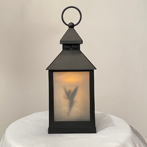 Könnte beinhalten: Schwarze Metalllaterne mit Griff und dekorativer Oberseite. Die Laterne hat eine Milchglasscheibe mit einer Silhouette einer Fee. Die Laterne steht auf einer weißen Oberfläche.