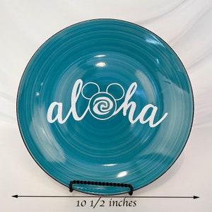 May include: A teal ceramic plate with a white "aloha" design, featuring a Mickey Mouse head in place of the "o". The plate has a spiral pattern and a black rim. It measures 10 1/2 inches in diameter.