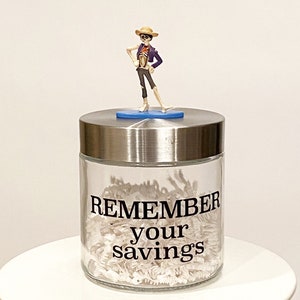 May include: A clear glass jar with a silver lid, filled with white shredded paper. The jar has the words "REMEMBER your savings" in black. A small figurine of a skeleton in a hat and purple jacket stands on top.