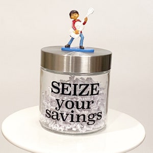 May include: A clear glass jar with a silver lid, filled with shredded paper. The jar has the words "SEIZE your savings" in black. A small figurine of a character playing a white guitar is on top.