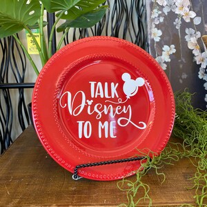 May include: A red decorative plate with a beaded edge and the phrase "Talk Disney To Me" in white script. A white Mickey Mouse balloon graphic is also featured. The plate is displayed on a black metal stand.