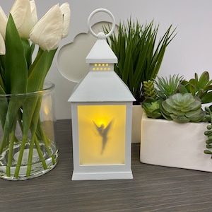 May include: A white metal lantern with a handle, featuring a silhouette of a fairy on the front panel. The lantern is illuminated from within, creating a soft glow. Decorative plants and flowers are in the background.
