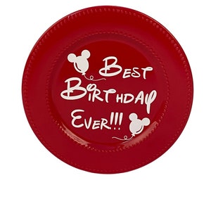 May include: A red ceramic plate with a beaded edge and the words "Best Birthday Ever!!!" in white script. Mickey Mouse head silhouettes are used as accents. A festive and celebratory design.