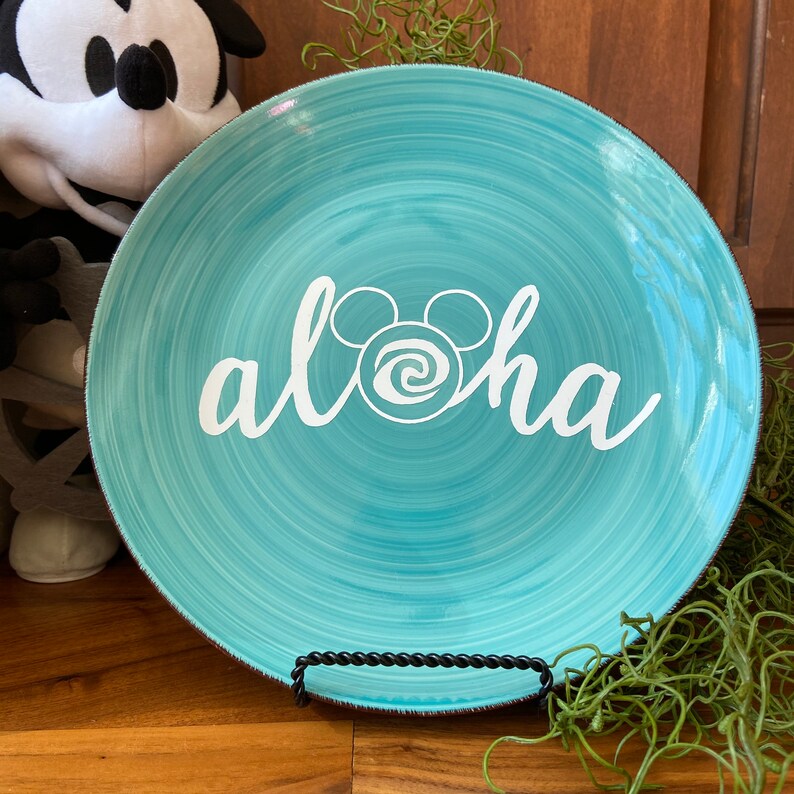 May include: A turquoise ceramic plate with a white "aloha" script and Mickey Mouse ears design. The plate has a swirled pattern and a dark rim. It is displayed on a black metal stand.