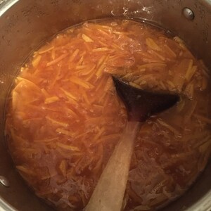 May include: A stainless steel pot filled with orange-colored marmalade, with a wooden spoon resting inside. The marmalade has a thick, glistening consistency, with visible pieces of orange peel. Steam is visible on the pot's interior.
