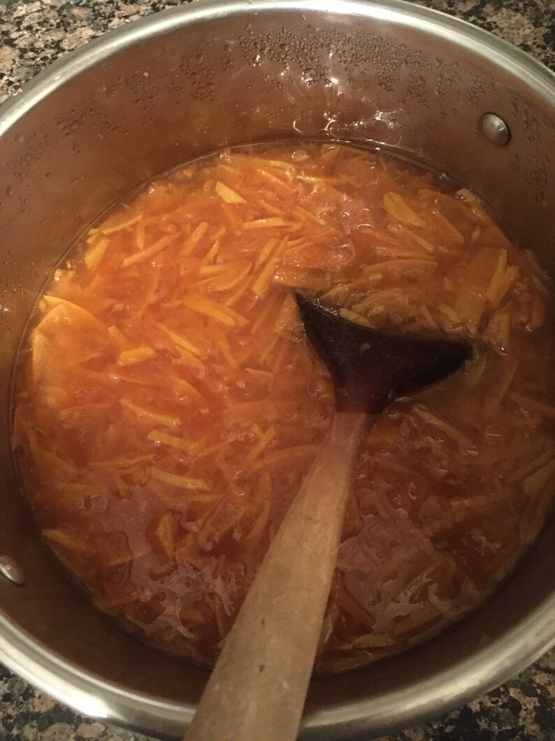 May include: A stainless steel pot filled with orange-colored marmalade, with a wooden spoon resting inside. The marmalade has a thick, glistening consistency, with visible pieces of orange peel. Steam is visible on the pot's interior.