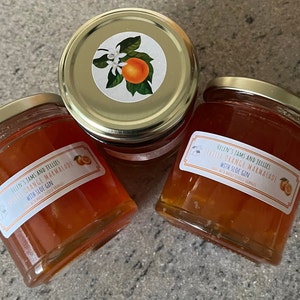 May include: Three glass jars of Seville Orange Marmalade with Sloe Gin. The jars have gold lids and labels with a white background and black text. The labels also feature a small orange and a sprig of orange blossoms.