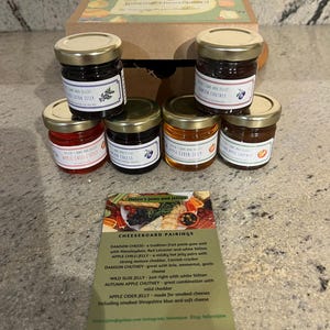May include: A set of six small glass jars filled with jams and jellies, arranged in a box. The jars have gold lids and labels with the text "Helen's Jams and Jellies". A card with the text "Cheeseboard Pairings" and pairing suggestions for each jam and jelly is in front of the jars.