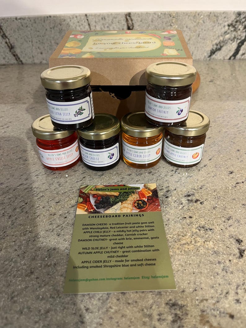 May include: A set of six small glass jars filled with jams and jellies, arranged in a box. The jars have gold lids and labels with the text "Helen's Jams and Jellies". A card with the text "Cheeseboard Pairings" and pairing suggestions for each jam and jelly is in front of the jars.