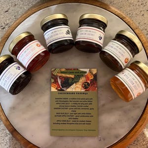 May include: A collection of six small glass jars filled with different jams and chutneys. The jars have gold lids and are arranged on a marble surface. The jars are labeled with the names of the jams and chutneys, including Damson Chutney, Apple Chilli Jelly, Wild Sloe Jelly, Autumn Apple Chutney, and Apple Cider Jelly. A card with the text "Helen's Jams and Jellies" and "Cheeseboard Pairings" is in the center of the arrangement.