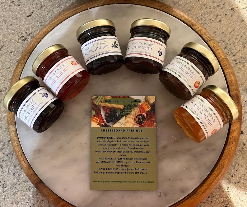 May include: A collection of six small glass jars filled with different jams and chutneys. The jars have gold lids and are arranged on a marble surface. The jars are labeled with the names of the jams and chutneys, including Damson Chutney, Apple Chilli Jelly, Wild Sloe Jelly, Autumn Apple Chutney, and Apple Cider Jelly. A card with the text "Helen's Jams and Jellies" and "Cheeseboard Pairings" is in the center of the arrangement.