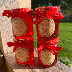May include: Four glass jars filled with bright red apple and rosehip jelly, stacked in two columns. Each jar features a red lid with white polka dots, a tied red ribbon, and a circular tan label with the handwritten text Apple Rosehip Jelly.