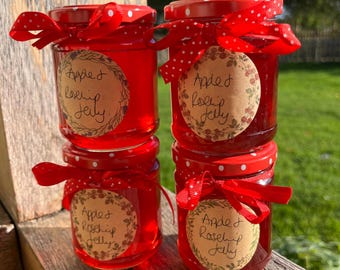 May include: Four glass jars filled with bright red apple and rosehip jelly, stacked in two columns. Each jar features a red lid with white polka dots, a tied red ribbon, and a circular tan label with the handwritten text Apple Rosehip Jelly.