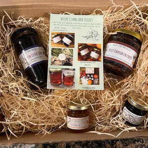 May include: A cardboard gift box filled with wood shavings containing four glass jars of preserves and a central informational card titled Helen's Jams and Jellies. The jars include Damson Extra Jam, Spicy Damson Chutney, and two smaller jars.