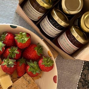 May include: A box of assorted jams and jellies with gold lids. The jams and jellies are labeled with the brand name "Melen's Jams and Jellies". The box is sitting on a table with a plate of strawberries, crackers, and cheese.