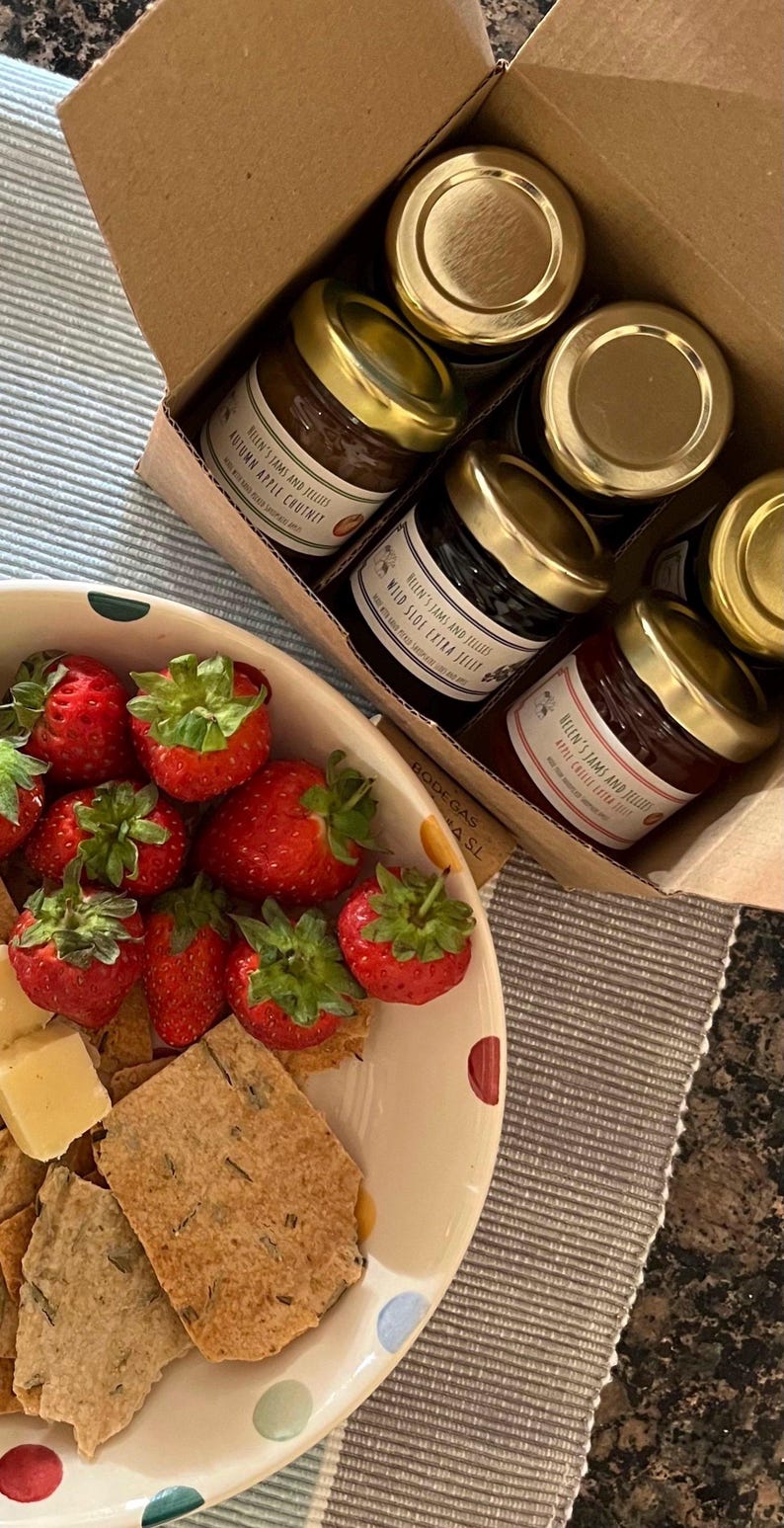 May include: A box of assorted jams and jellies with gold lids. The jams and jellies are labeled with the brand name "Melen's Jams and Jellies". The box is sitting on a table with a plate of strawberries, crackers, and cheese.