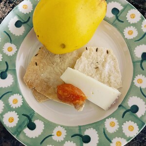May include: A green and white plate with a daisy pattern, featuring a yellow quince, two pieces of bread, a slice of cheese, and a small dish of orange jam.