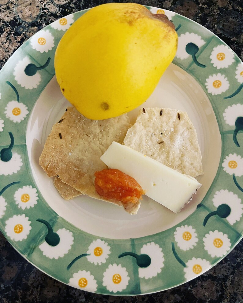 May include: A green and white plate with a daisy pattern, featuring a yellow quince, two pieces of bread, a slice of cheese, and a small dish of orange jam.