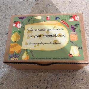May include: A brown cardboard box with a colorful label that says "Homemade condiments for your cheeseboard" and "6 mini jars for your cheeseboard". The label features a cartoon illustration of a cheese wedge with various fruits, vegetables, and herbs.