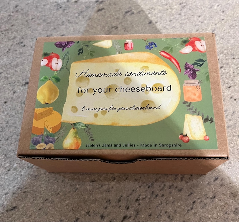 May include: A brown cardboard box with a colorful label that says "Homemade condiments for your cheeseboard" and "6 mini jars for your cheeseboard". The label features a cartoon illustration of a cheese wedge with various fruits, vegetables, and herbs.