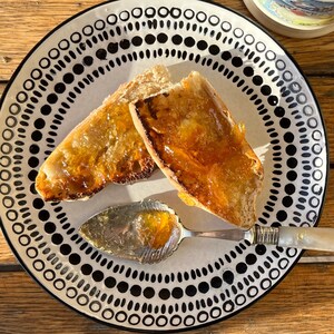 May include: A white plate with a black dotted pattern holds two slices of toasted bread topped with orange marmalade. A silver serving spoon with a mother-of-pearl handle rests on the plate, with a small amount of marmalade.