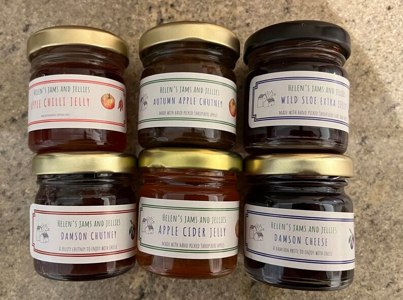 May include: Six small glass jars with gold lids filled with different jams and jellies. The labels are white with black text and feature a small illustration of a house. The labels read: Apple Chilli Jelly, Autumn Apple Chutney, Wild Sloe Extra Jelly, Damson Chutney, Apple Cider Jelly, and Damson Cheese.