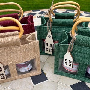 May include: Four small rectangular burlap gift bags with rounded wooden handles. The bags come in tan, burgundy, and two shades of green. Each bag features a clear plastic window at the bottom and a small wooden house-shaped gift tag.