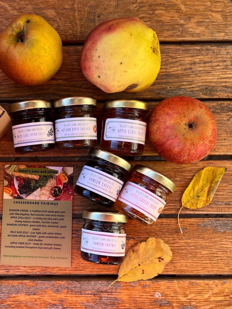 May include: Assortment of small glass jars filled with jams and jellies, including Damson Cheese, Apple Chilli Jelly, Wild Sloe Jelly, Autumn Apple Chutney, and Apple Cider Jelly. The jars have gold lids and labels with the name "Helen's Jams and Jellies". There are also apples and fall leaves on a wooden surface.