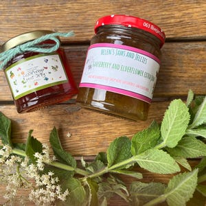 May include: Two glass jars of jam with gold lids and labels. One jar is tied with a teal ribbon. The labels read "Gooseberry Mint Jelly" and "Helen's Jams and Jellies Gooseberry and Elderflower Extra Jam". Fresh green leaves and white flowers are in the foreground.