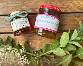 May include: Two glass jars of jelly with gold lids and labels. One jar is tied with a teal ribbon. The labels read "Gooseberry Mint Jelly" and "Helen's Jams and Jellies Gooseberry and Elderflower Extra Jam". Fresh green leaves and white flowers are in the foreground.