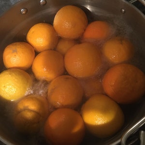 May include: A stainless steel pot filled with oranges in boiling water. The oranges vary in color from bright orange to yellow. Steam rises from the water, indicating the oranges are being cooked. The pot's handle is visible on the right side.