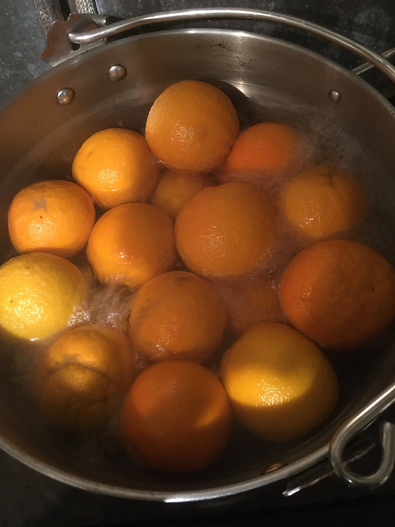 May include: A stainless steel pot filled with oranges in boiling water. The oranges vary in color from bright orange to yellow. Steam rises from the water, indicating the oranges are being cooked. The pot's handle is visible on the right side.