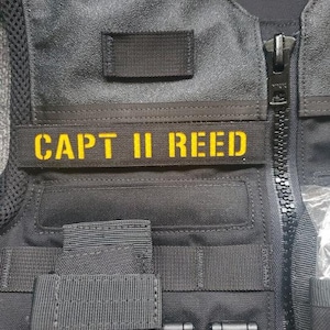Peut inclure: Gros plan sur un gilet tactique noir avec un écusson brodé jaune portant l'inscription "CAPT II REED". Le gilet est doté d'une fermeture éclair, de multiples sangles et d'un petit écusson carré au-dessus du nom. Le gilet semble être fait d'un matériau durable et robuste.