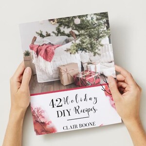 以下が含まれることがあります： 「42 Holiday DIY Recipes」というタイトルのレシピ本を人が持っています。Clair Boone著。表紙には赤い毛布、プレゼント、クリスマスツリーが入ったバスケットが描かれています。