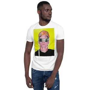 May include: A white t-shirt featuring a colourful illustration of a person wearing sunglasses and a helmet with the word "DEVO" on it. The illustration is set against a yellow background.