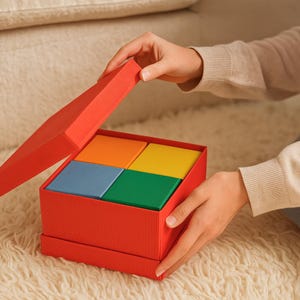 May include: A red gift box with a textured exterior is being opened to reveal four colorful squares: orange, yellow, blue, and green. The box is held by a person, and the background is a soft, neutral color.