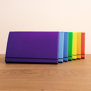 May include: A row of colorful, corrugated paper document holders in purple, blue, green, yellow, and red. Each folder has a black elastic band closure. The folders are arranged on a wooden surface.
